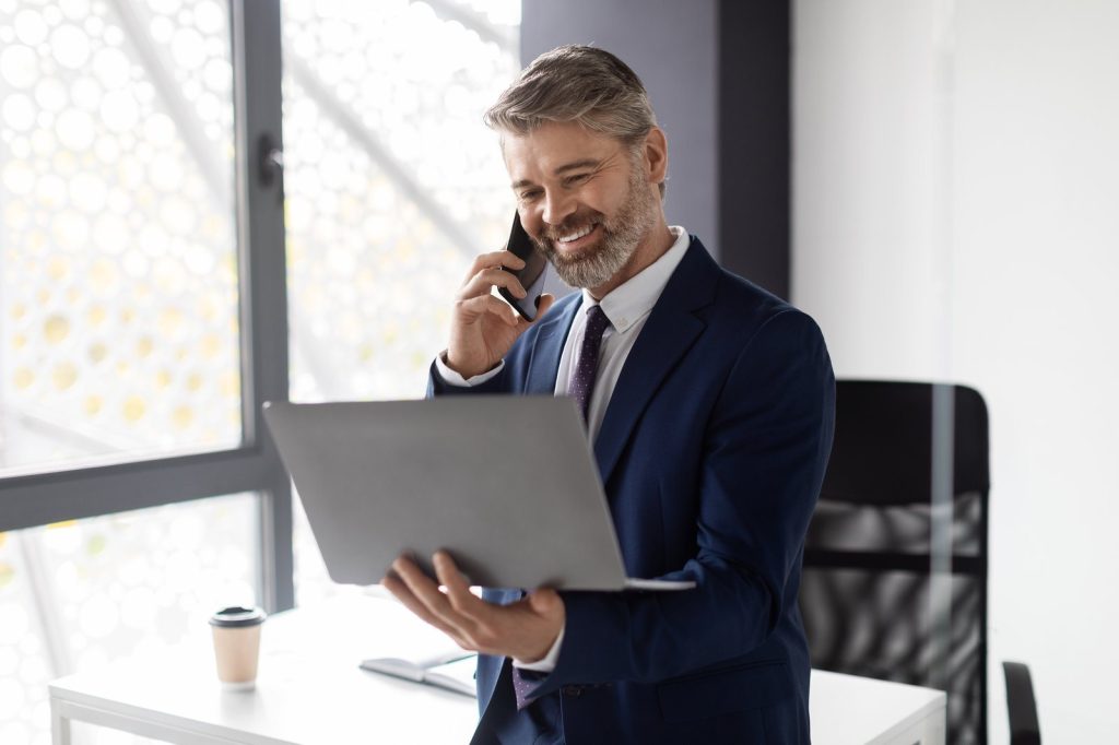 Smiling adviser holding a laptop while talking on the phone by an office window