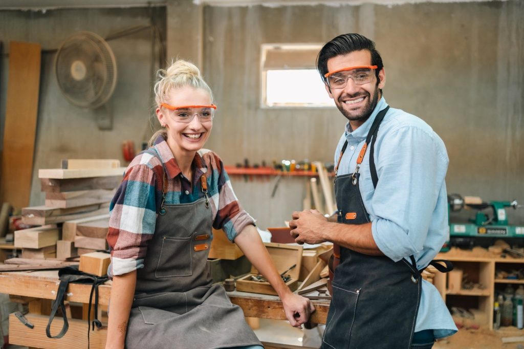 Two smiling woodworkers in safety goggles standing in their workshop