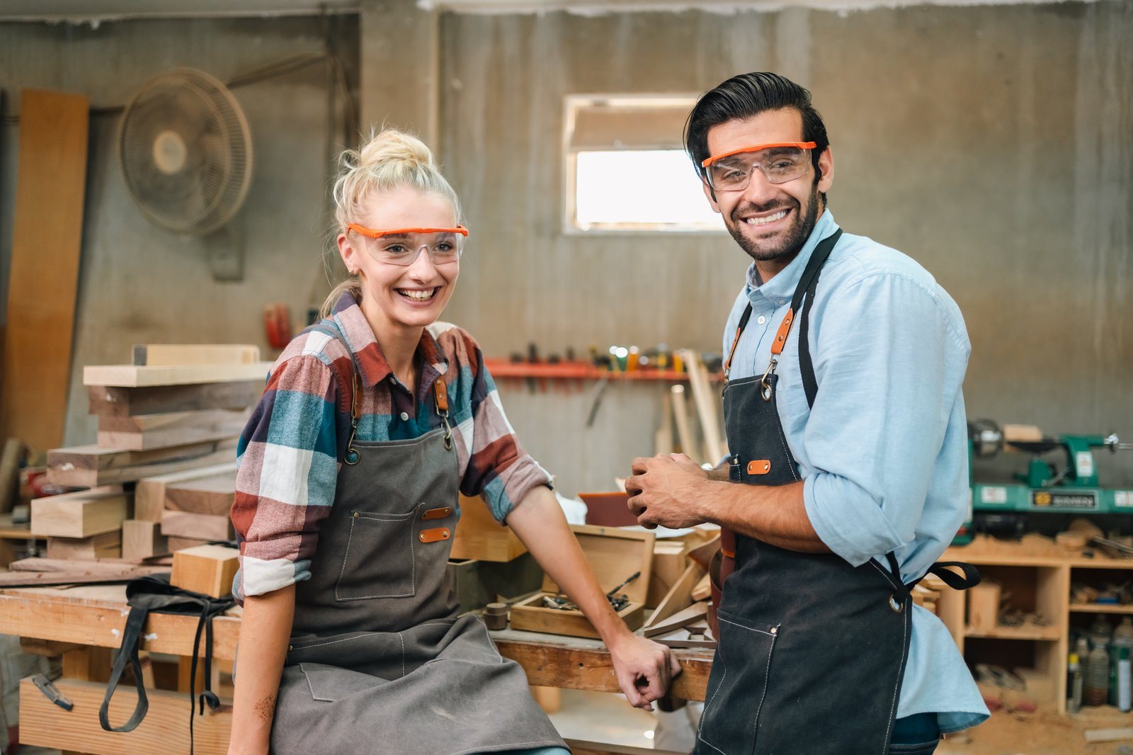 Two smiling woodworkers in safety goggles standing in their workshop