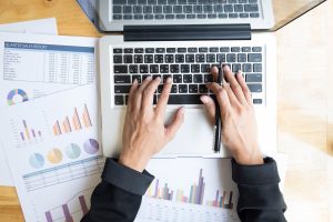 Hands typing on a laptop surrounded by printed business charts and reports