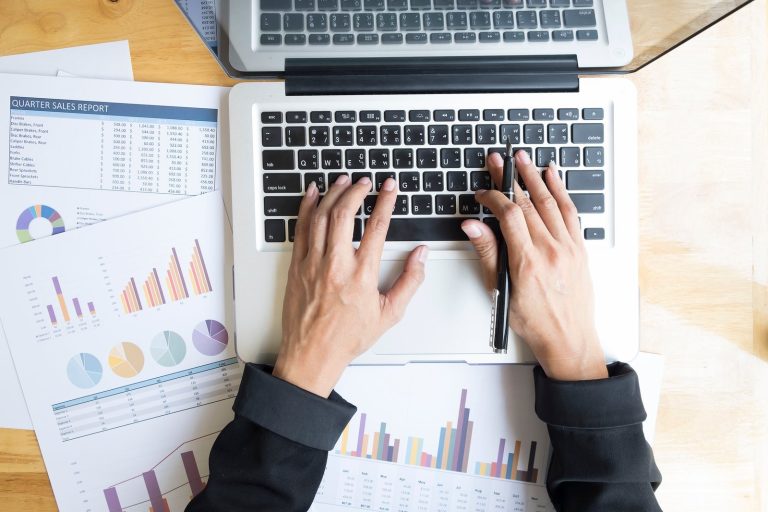 Hands typing on a laptop surrounded by printed business charts and reports